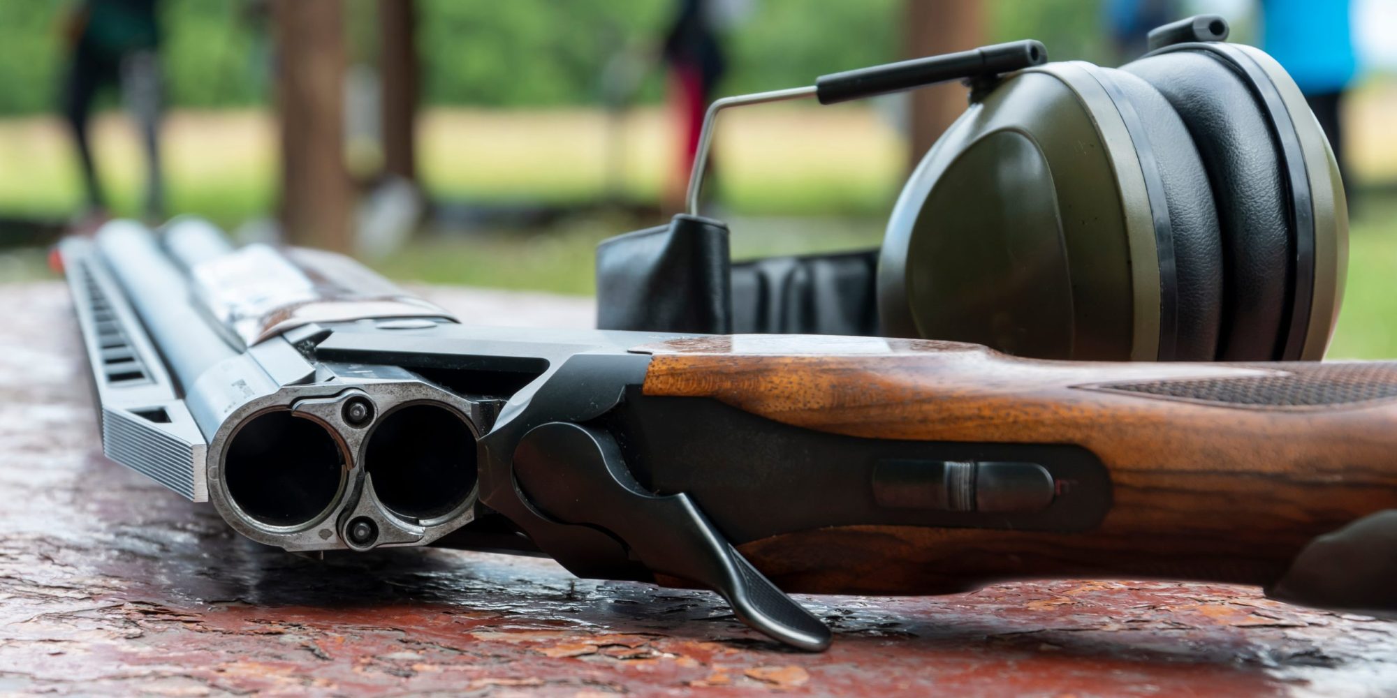 A sports double-barreled shotgun and headphones for shooting lie on a wooden table against the background of the shooting range.